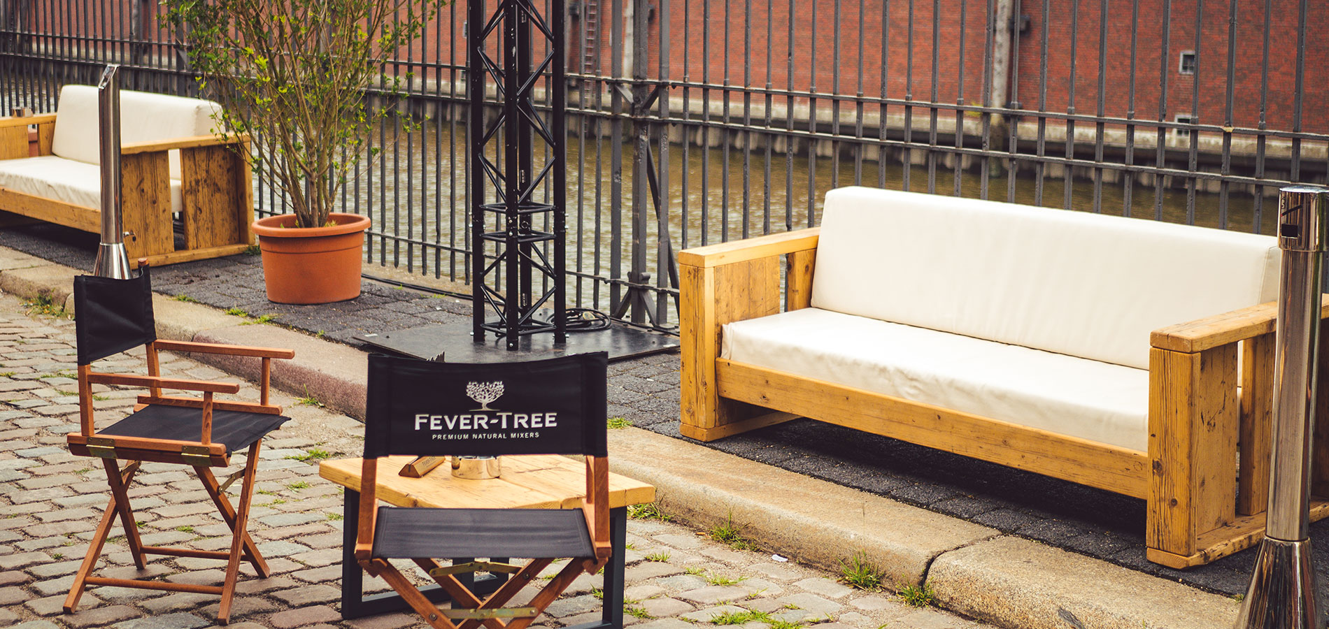 Outdoor riverside seating area with white-cushioned wooden sofas and folding director chairs by a brick building and iron fence, Fever-Tree-branded chair in foreground.